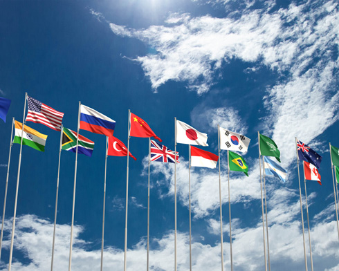 A row of international flags representing various countries, waving against a bright blue sky with scattered clouds.